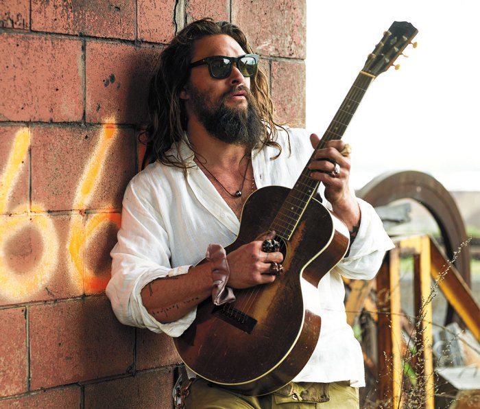 Jason Momoa wearing sunglasses and white button-down shirt playing guitar