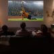 Three men sitting on couch watching soccer game off projector