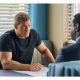Actor Alan Ritchson sits at a table in a muscle-bound t-shirt during a scene from Amazon's "Reacher"