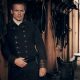 'Outlander' star Sam Heughan stands beside a horse stable wall in character in a dark double breasted jacket