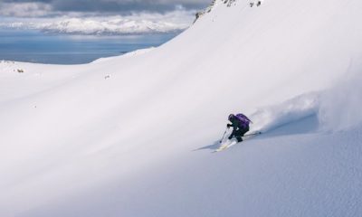 Skier heads down a slope of powder in Northern Norway with coastal waters in the far background.