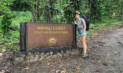 Woman in a backpack and hiking shorts standing next to a Na Pali Coast State Park sign near a muddy path. Thru-hike