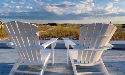 A pair of empty white Adirondack chairs on a porch in Cape Cod overlooking the water.