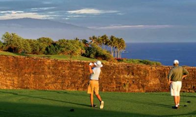 Two golfers, one taking a swing, at a golf course tee box overlooking a cliff. Golfing in Hawaii