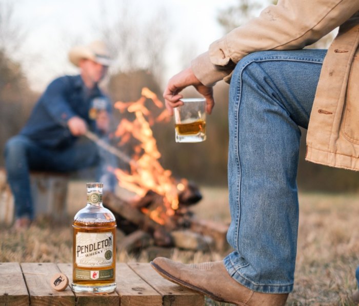 Man out of focus stoking fire in background while man in foreground has leg on table with bottle of Pendleton Whisky