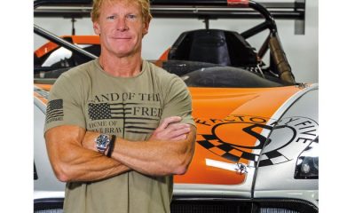 Caucasian man with strawberry blonde hair poses with arms across chest in front of sports car